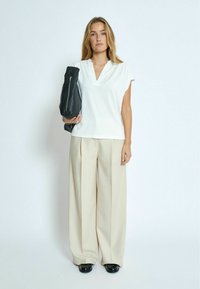 Blusa bianca a scollo a V con maniche corte, abbinata a pantaloni larghi color beige chiaro e scarpe nere, mentre tiene in mano una borsa in pelle nera.