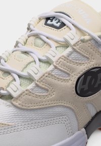 Κοντινό πλάνο ενός μπεζ και λευκού αθλητικού παπουτσιού με λευκά κορδόνια, επενδύσεις από σουέντ, ύφασμα από δίχτυ και λογότυπο DC στο πλάι.