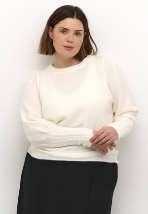 Cream-colored knitted sweater with a round neck and ribbed cuffs, featuring a soft texture and relaxed fit. Paired with a black skirt.