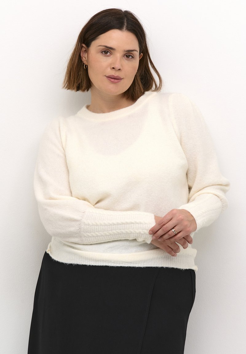 Pull en tricot couleur crème avec un col rond et des poignets côtelés, présentant une texture douce et une coupe décontractée. Associé à une jupe noire.