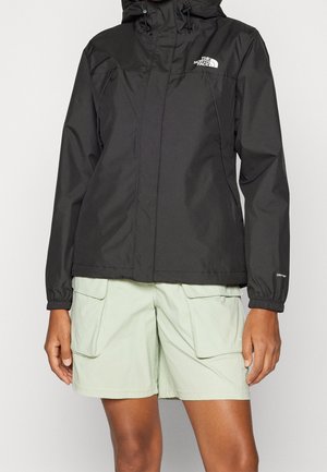 Person, die eine schwarze North Face Kapuzenjacke mit elastischen Bündchen und hellgrüne Cargoshorts mit großen Klappentaschen trägt.
