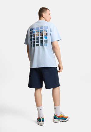 Mężczyzna stoi tyłem, nosząc jasnoniebieską koszulkę z grafiką siatki zdjęć na plecach, granatowe szorty, białe skarpetki i kolorowe trampki.