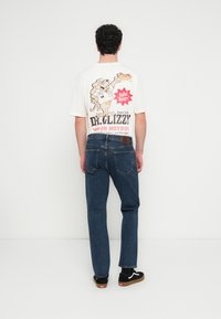 Άνδρας που φοράει λευκό μπλουζ άκι με σχέδιο καρτούν σκύλου που κρατάει χοτ ντογκ και κείμενο, συνδυασμένο με μπλε τζιν και μαύρα αθλητικά παπούτσια, στέκεται κοιτώντας έναν τοίχο.