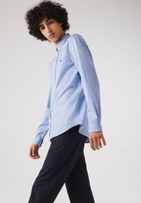 Camicia a quadri blu e bianchi con colletto a punta, maniche lunghe e un logo sottile; abbinata a pantaloni scuri.