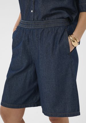 Personne portant un short en denim bleu foncé et un haut boutonné assorti, les mains dans les poches et des bracelets dorés au poignet.