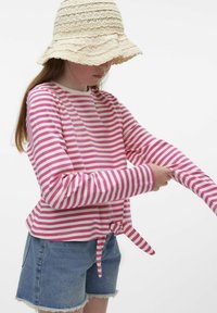Camicia a maniche lunghe con strisce rosa e bianche, caratterizzata da un dettaglio in vita annodato. Abbinata a pantaloni corti di jeans, il modello indossa un cappello intrecciato beige.
