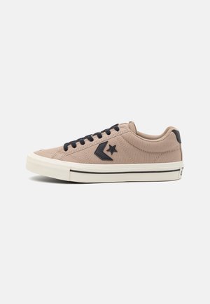 Beżowy niski sneaker z czarnymi sznurowadłami, czarną gwiazdą i logo w kształcie chevronu oraz białą podeszwą z czarnym paskiem.