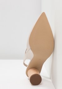 Chaussure à talon haut beige à bout pointu avec un talon bloc en bois rond, vue de la semelle reposant contre un mur blanc.
