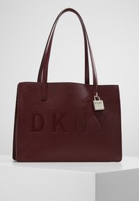 Burgundowa skórzana torba typu tote z teksturowaną powierzchnią, z wytłoczonym logiem "DKNY" i metalową zawieszką z logo przymocowaną do uchwytu.
