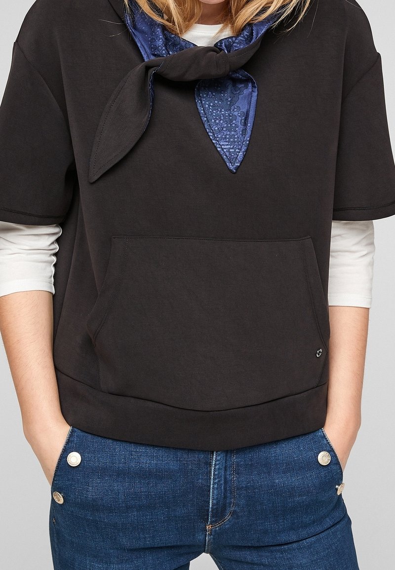 Personne portant des vêtements superposés : un sweat-shirt noir à manches courtes sur une chemise blanche à manches longues, un foulard bleu à motifs autour du cou et un jean bleu avec des boutons argentés.