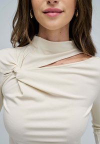 Camiseta de manga larga en color crema con un cuello alto, que presenta un detalle de nudo retorcido y un corte abierto en un lado. Tejido suave y elástico.