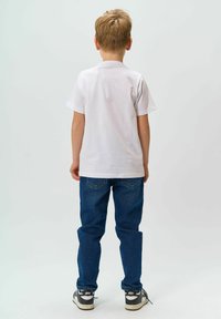 Biała koszulka z krótkim rękawem, niebieskie dżinsy i czarne sneakersy. Spodnie wykonane są z denimu w klasycznym pięciokieszeniowym wzornictwie i prostym kroju.