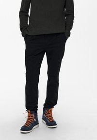 Schwarze, zulaufende Hose kombiniert mit dunkelblauen Wanderschuhen mit orangen Schnürsenkeln und silbernen Beschlägen. Das Oberteil ist ein dunkel gerippter Pullover.