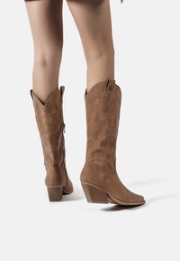 Piernas con botas vaqueras marrones de media caña con patrones decorativos perforados y tacones apilados sobre un fondo blanco.