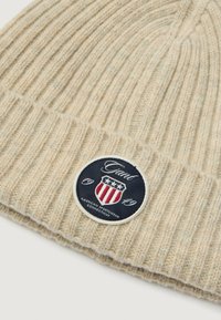 Gorro de punto acanalado beige hecho de una tela suave. Presenta un parche negro redondo con un diseño de escudo, estrellas y el texto "Gant 1949."