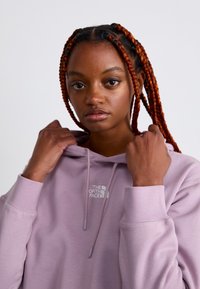 Sweatshirt violet clair avec capuche, présentant un petit logo blanc sur la poitrine et des poignets côtelés. Texture lisse et coupe décontractée.