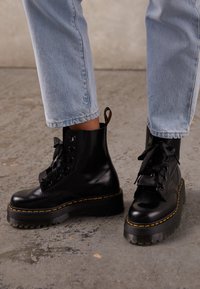 Botas de tobillo de cuero negro con suela gruesa y texturizada, costuras amarillas y acabado brillante; con diseño de cordones y asa en bucle.