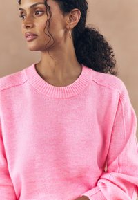 Suéter rosa de punto con cuello redondo acanalado, con hombros caídos y textura suave. Destacan los puños enrollados y un ajuste relajado.