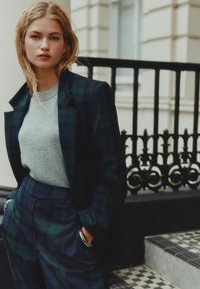 Sacou la comandă din tartan verde închis cu rever ascuțite, combinat cu un pulover gri deschis și pantaloni cu tartan asortați. Material neted și croială structurantă.