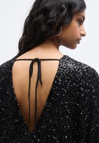 Μαύρο φόρεμα με παγιέτες και βαθύ V στο πίσω μέρος, δεμένο με λεπτή μαύρη κορδέλα. Διαθέτει μικρά μανίκια και λαμπερή υφή από παγιέτες.