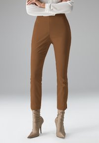 Braune, hoch taillierte Hosen mit glatter Textur, schmal zulaufender Form und Knöchellänge, kombiniert mit beigen kniehohen, eng anliegenden Stiefeln mit Absatz.