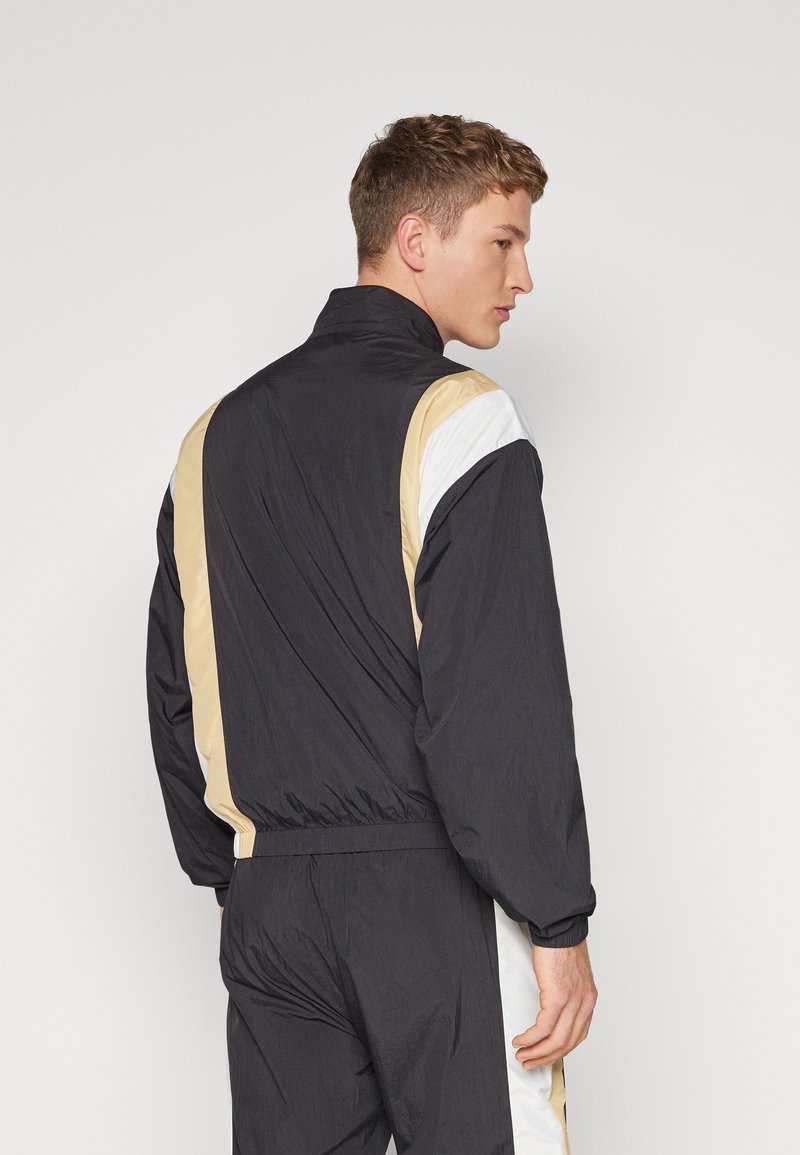 Veste coupe-vent noire avec des accents beige et blanc sur les épaules, présentant un col haut et des poignets élastiques. Tissu léger avec une texture lisse.