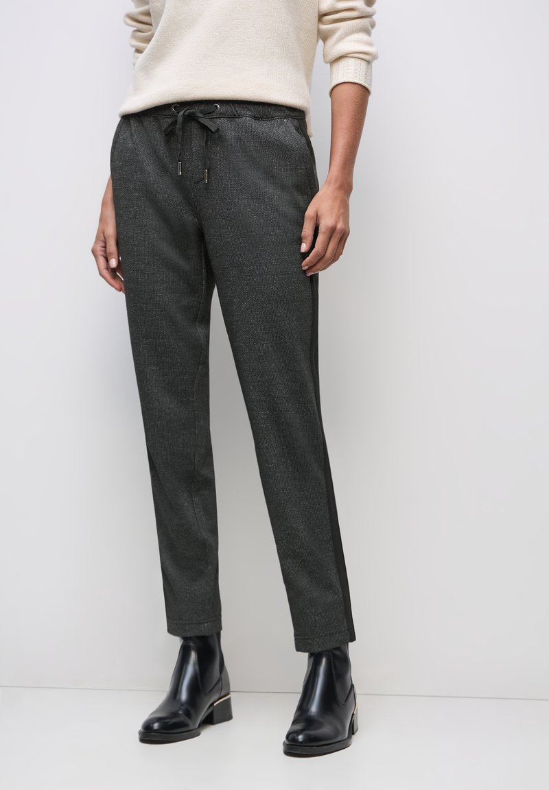 Femme portant un pantalon gris foncé à cordon avec des rayures latérales noires et des bottines noires, debout devant un fond clair uni.