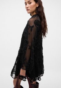 Crna prozirna haljina s cvjetnim vezom, duge rukave, visoki ovratnik, lepršavi oblik, uparen sa burgundskom torbicom.