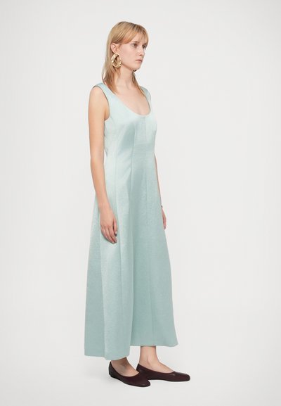Un vestito senza maniche di colore verde menta con scollatura a barca e orlo svasato, realizzato in tessuto testurizzato, abbinato a ballerine scure.