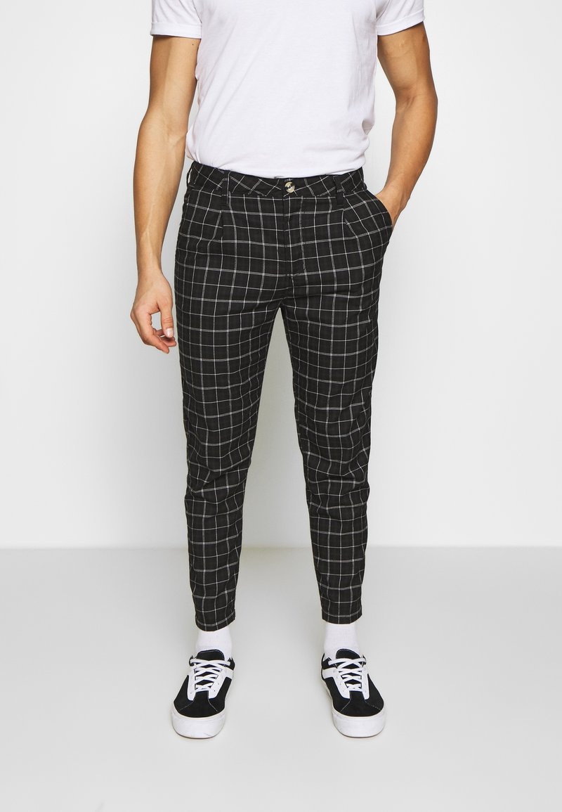 Homme portant un pantalon slim à carreaux noir et blanc, des chaussettes blanches, des baskets noir et blanc, et un t-shirt blanc uni, debout avec une main dans la poche.