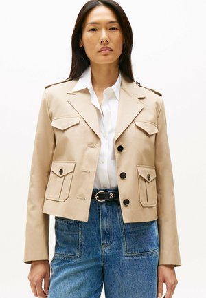 Beige cropped jack met vier voorzakken, zwarte knopen en epauletten. Draagt over een wit shirt, gecombineerd met high-waisted denim.