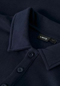 Polo de color azul marino oscuro con tres botones y un cuello doblado, mostrando una etiqueta de ropa negra con texto blanco en el interior del cuello.