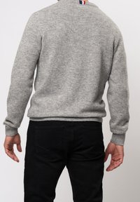 Grå stickad tröja med ribbade muddar och nederkant. Har en liten randig halsringning i röd, vit och blå. Bärs med svarta byxor.