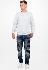 Sweatshirt gris clair à col rond avec manches longues, associé à un jean cargo bleu déchiré avec de multiples poches zippées et des accents noirs. Baskets blanches.