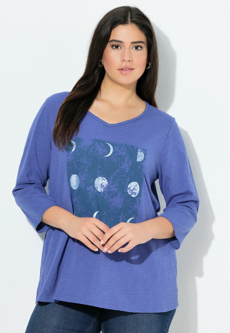 Vrouw met lang donker haar, gekleed in een blauwe shirt met driekwart mouwen en maanfase graphics, staande tegen een witte achtergrond.