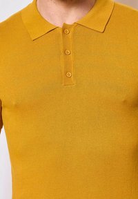 Polo shirt jaune avec une texture côtelée, présentant un col et trois boutons au niveau de l'encolure. La coupe est ajustée au corps.