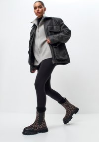 Stivali alla caviglia con stampa leopardata, suola in gomma nera e polsini a coste, abbinati a un maglione grigio e leggings neri, su uno sfondo bianco.