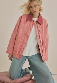 Rosa denimjakke med en fløjlskrave, flere frontlommer og sølvhardware, båret over en hvid t-shirt og blå jeans.
