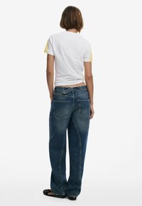 Lösa blå denimjeans med en blekt tvätt, har två bakfickor och dragkedja. Matchas med en vit t-shirt med gula detaljer.