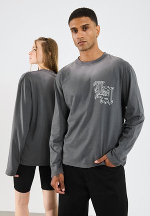 Langermet grå t-skjorte med "LA" logo på forsiden, laget av mykt stoff. Modellen er kombinert med svarte shorts, minimal tekstur og enkelt design.