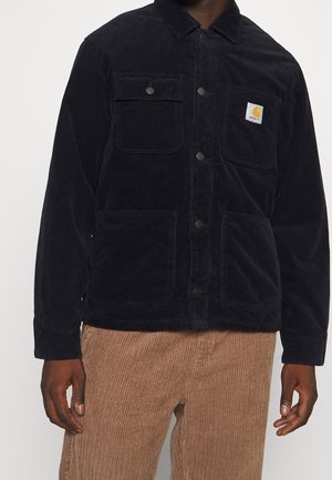 Mand iført sort corduroy Carhartt-jakke med frontklap og lappelommer, kombineret med lysebrune corduroy-bukser.