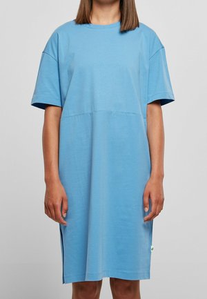 Frau in einem locker sitzenden, kurzärmligen, hellblauen knielangen Kleid mit seitlichen Schlitzen vor einem schlichten grauen Hintergrund.