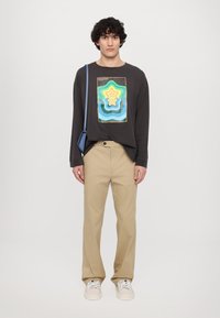 Čierna košeľa s dlhým rukávom so farebnou grafikou hviezdy, v kombinácii s béžovými nohavicami a bielymi teniskami. Obsahuje modrú cez rameno nosenú tašku.