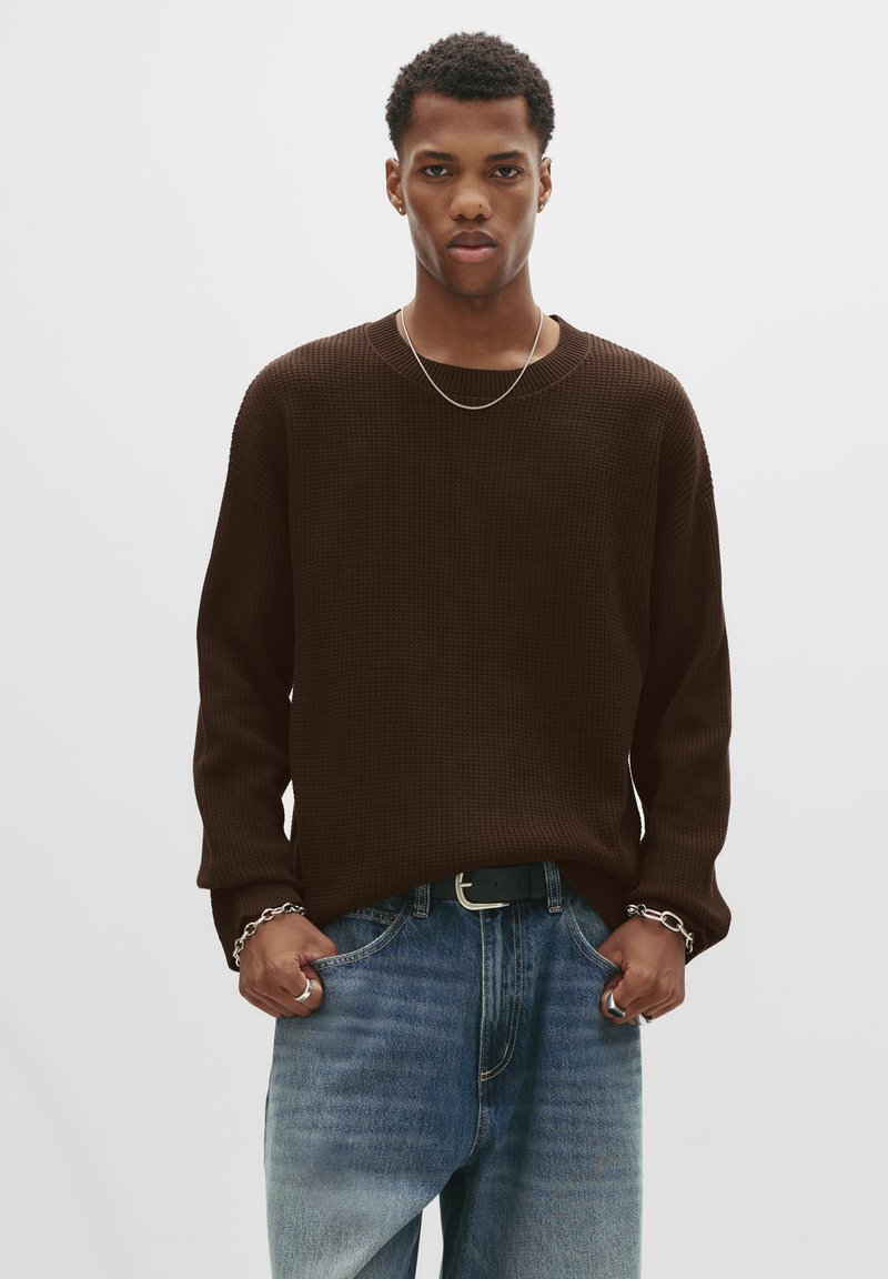 Homme portant un pull marron foncé, un jean bleu, un collier en chaîne argentée, un bracelet et des bagues, se tenant avec les mains dans les poches sur un fond blanc uni.