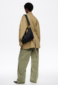 Melnas neilona plecu somas ar izliekta formu, kurai ir augšējais rokturis un regulējama siksna. Modelis valkā pārāk lielu beigu žaketi un zaļas bikses.