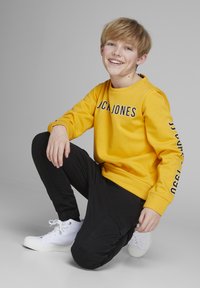 Yellow sweatshirt with "JCJ JONES" text, black pants with side pockets, and white sneakers. Smooth texture, casual fit, and relaxed posture.