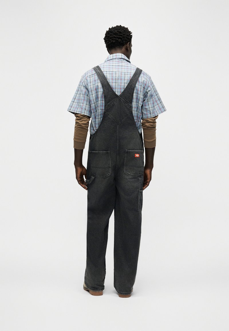 Uomo che indossa una salopette di denim nera sopra una maglietta marrone a maniche lunghe e una camicia a quadri a maniche corte, in piedi di spalle su uno sfondo semplice.