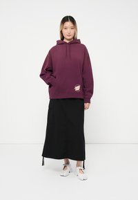 Felpa con cappuccio bordeaux con tasca frontale e logo, abbinata a una gonna nera alla caviglia e scarpe slip-on grigie.
