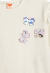 Pull en coton crème avec trois patchs décoratifs de papillons en sequins et broderies, présentant des couleurs et motifs variés.