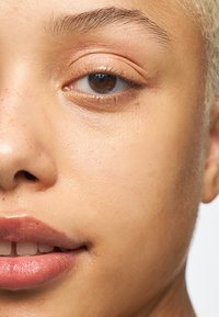 Gros plan d'un visage à la peau claire avec des taches de rousseur, montrant un œil marron, des lèvres pleines avec un éclat naturel et des cheveux courts blonds plats.